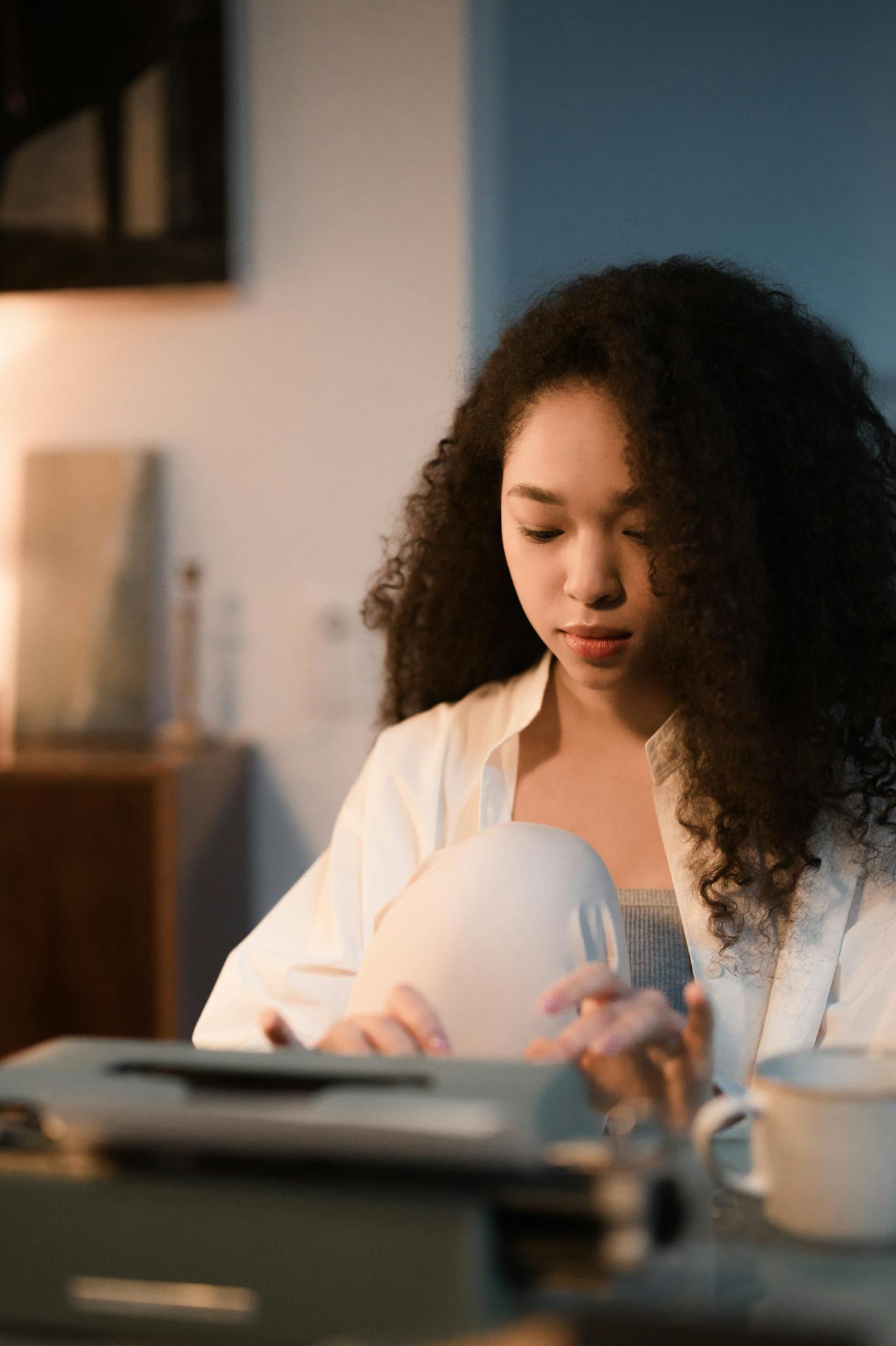 A focused woman with curly hair types on a typewriter in a warmly lit indoor setting, creating content.
